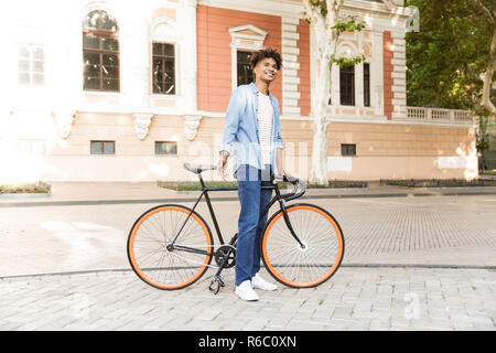 Immagine di bello emotivo giovane ragazzo africano sulla strada all'aperto camminando con la bicicletta. Foto Stock