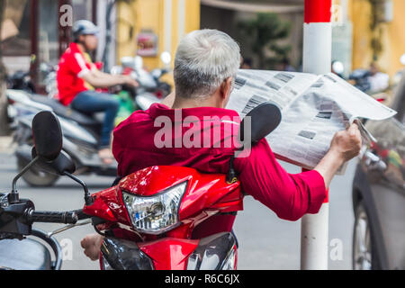 Irriconoscibile Uomo in camicia rossa si legge il giornale, si siede sullo scooter, il traffico in background, Hanoi Vietnam Foto Stock