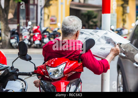 Irriconoscibile Uomo in camicia rossa si legge il giornale, si siede sullo scooter, vuoto Street di Hanoi, Vietnam Foto Stock