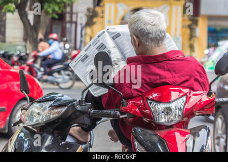 Irriconoscibile Uomo in camicia rossa si legge il giornale, si siede sullo scooter, il traffico in background, Hanoi Vietnam Foto Stock