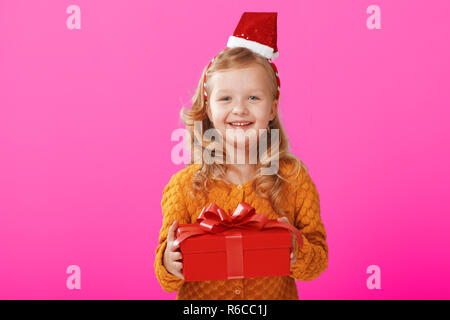 Felice piccolo bambino ragazza in un maglione caldo e cappuccio Babbo Natale è in possesso di una casella rossa con un regalo su uno sfondo di colore rosa. Il concetto di celebratio Foto Stock