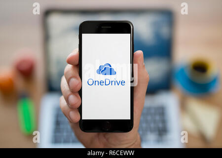Un uomo guarda al suo iPhone che visualizza il Microsoft logo OneDrive, mentre se ne sta seduto alla sua scrivania per computer (solo uso editoriale). Foto Stock