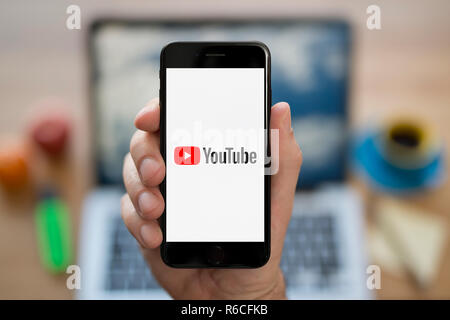 Un uomo guarda al suo iPhone che visualizza il logo di Youtube, mentre se ne sta seduto alla sua scrivania per computer (solo uso editoriale). Foto Stock