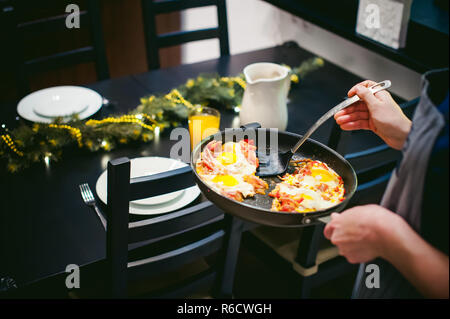 Donna mette cibo fatto in casa. Mani femminili applicato con cautela per uovo fritto piatto dal pan. La prima colazione per due persone a casa in cucina al tavolo da pranzo Foto Stock