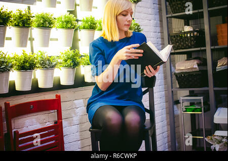 Donna bionda con un vestito si siede su una sedia con un libro in mano, contro lo sfondo di houseplants. impegnato nella lettura Foto Stock