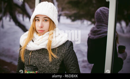 La donna in bianco, sciarpa e cappello beve caffè. cute giovane donna bionda in piedi vicino a specchio vetrine cafe, bere il caffè per andare, tenendo il Natale bicchiere di carta nelle sue mani Foto Stock