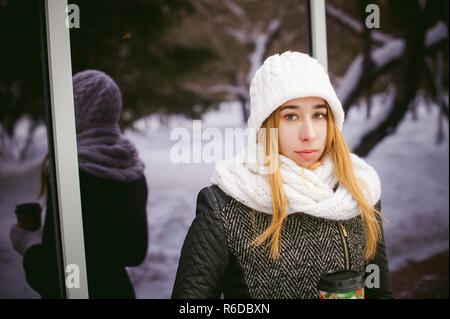 La donna in bianco, sciarpa e cappello beve caffè. cute giovane donna bionda in piedi vicino a specchio vetrine cafe, bere il caffè per andare, tenendo il Natale bicchiere di carta nelle sue mani Foto Stock