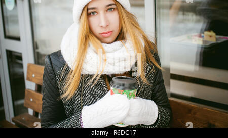 La donna in bianco, sciarpa e cappello beve caffè. cute giovane ragazza bionda seduta su una panchina vicino alla caffetteria, bere il caffè da asporto, tenendo il Natale bicchiere di carta nelle sue mani. guardando la telecamera Foto Stock