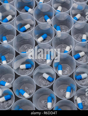 Big pharma farmaco concetto. Blu e bianco capsule pillole nel bicchiere graduato in plastica / coppa di erogazione sulla superficie riflettente. Possibile metafora di NHS. Foto Stock