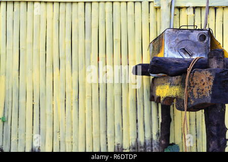 Quindici litri di piazza vecchia olio da cucina in metallo può svuotare e rusty collocato su un tavolo di legno sulla sabbia della spiaggia-dipinte di un giallo pannello di bambù o di recinzione Foto Stock
