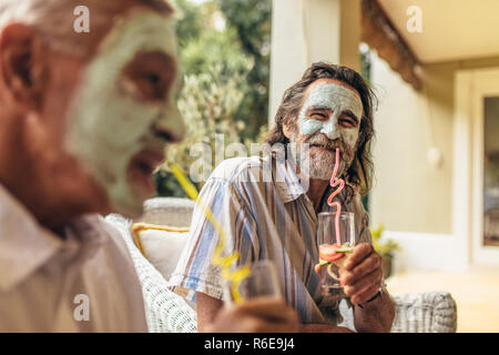 Due amici senior con il facciale della maschera di argilla sul bere il succo. Pensionati uomini seduti insieme con la maschera per il viso con il succo. Foto Stock