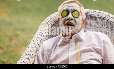 Ritirato uomo seduto su una sedia con argilla maschera al viso e Cucumber Slice sulla faccia. Senior uomo prendendo facial trattamento termale. Foto Stock