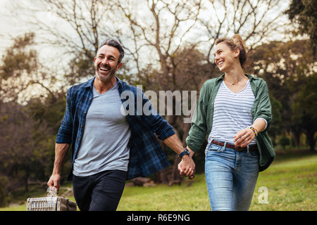 Coppia felice passeggiate nel parco tenendo la mano e portando un cestino picnic. Amare giovane andare a fare un picnic al parco. Foto Stock