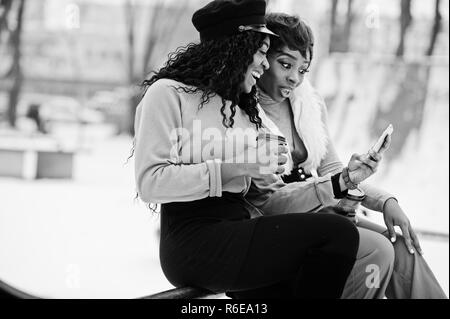 Due americano africano womans poste a giornata invernale contro sfondo innevato con tazze di caffè e guardando sul telefono. Foto Stock