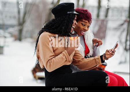 Due americano africano womans poste a giornata invernale contro sfondo innevato con tazze di caffè e guardando sul telefono. Foto Stock