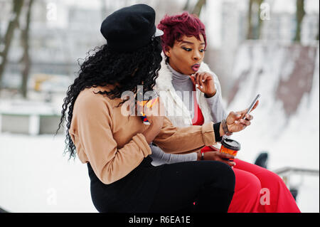 Due americano africano womans poste a giornata invernale contro sfondo innevato con tazze di caffè e guardando sul telefono. Foto Stock