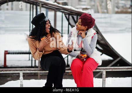 Due americano africano womans poste a giornata invernale contro sfondo innevato con tazze di caffè e guardando sul telefono. Foto Stock