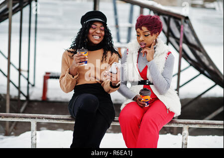 Due americano africano womans poste a giornata invernale contro sfondo innevato con tazze di caffè e guardando sul telefono. Foto Stock