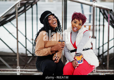 Due americano africano womans poste a giornata invernale contro sfondo innevato con tazze di caffè. Foto Stock