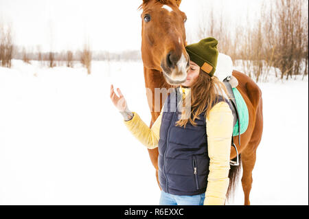 Pilota femmina e il cavallo in aria aperta. ritratto di una giovane e bella donna con il suo stallone, all'aperto in inverno. ragazza abbracciando e accarezzando l'animale Foto Stock