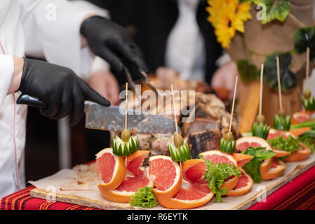 Il capo cuoco tagli in pezzi cucinata fresca di maiale hip, processo close up. Decorazioni di frutta e verdura sul tavolo, catering. Spettacolo al ristorante Foto Stock