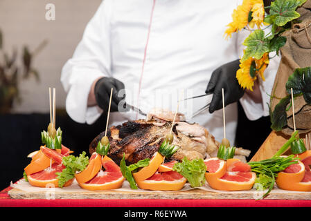 Spettacolo al ristorante: il capo cuoco tagli in pezzi appena cucinata coscia di maiale, il processo da vicino. Decorazioni di frutta e verdura sul tavolo, catering Foto Stock