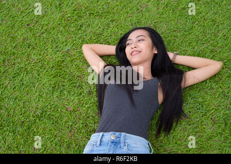 Felice giovane bella donna asiatica che stabilisce su erba presso il parco Foto Stock