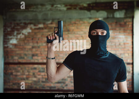 Athletic uomo in un passamontagna, maschera da sci sulla sua testa, detiene una pistola nella sua mano, sollevando una canna, un gangster pericolose Foto Stock