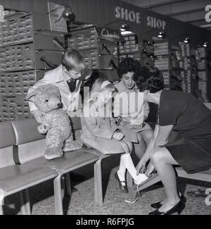 Anni '1960, storica, due bambini con la madre all'interno di un negozio di scarpe, la giovane ragazza seduta su una sedia che misura i piedi da un'assistente di negozio, utilizzando una misura tradizionale del piede, Inghilterra, Regno Unito. Foto Stock