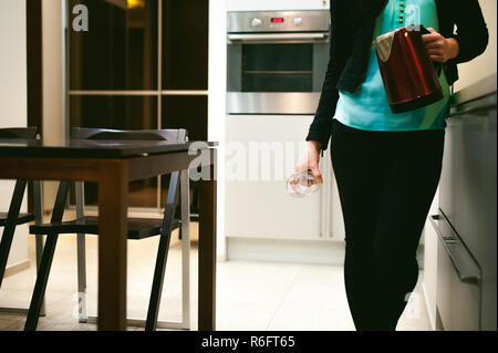 Donna bionda in cucina tenendo un bicchiere di vetro, versare l'acqua dal bollitore per soddisfare la loro sete Foto Stock