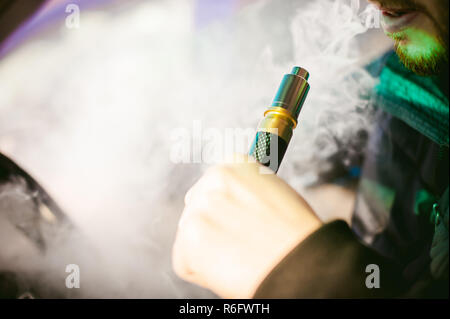 Giovane uomo vaper con la barba vaping meccanica mod. Guy fuma una sigaretta elettronica mediante insufflazione di vapore di fumo. Tiene in mano su sfondo colorato Foto Stock