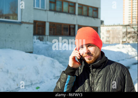 L'uomo emotivamente comunica telefonicamente. guy con la barba, vestito di un autunno sport giacca casual e arancione brillante hat, parla da telefono cellulare in elegante custodia, passeggiate all'aperto sulla soleggiata giornata invernale Foto Stock