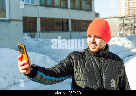 Uomo di fotografare sé. guy con la barba, vestito di un autunno sport giacca casual e arancione brillante hat, rende selfie sul telefono in elegante custodia, passeggiate all'aperto sulla soleggiata giornata invernale Foto Stock