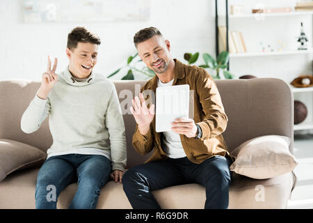Padre e figlio teen agitando la mano e mostrando segno di pace durante la chat video a casa Foto Stock