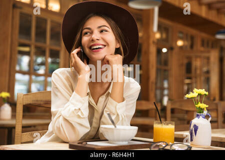 Ritratto di donna rilassata indossando hat parlando sullo smartphone mentre è seduto in intimo cafe Foto Stock