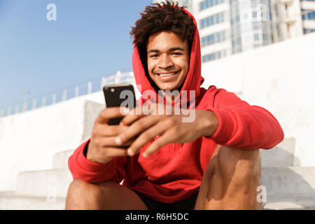 Immagine di un giovane ragazzo all'aperto sulla spiaggia seduti sui gradini sorridente utilizzando il telefono cellulare. Foto Stock