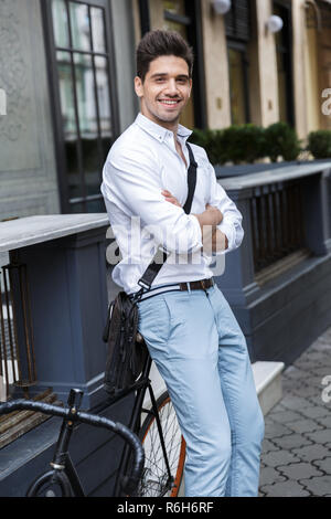 Imprenditore sorridente vestito in camicia in piedi all'aperto presso la via della città portando un sacco, appoggiato su di una bicicletta Foto Stock