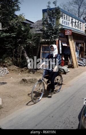 Una donna che indossa abbigliamento tradizionale giapponese cavalca una bicicletta rapido su una strada sterrata in passato un negozio con segni nel tradizionale giapponese, script in Giappone, 1955. () Foto Stock