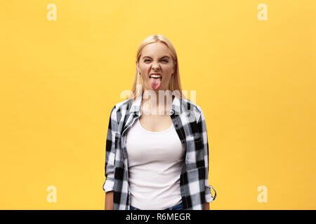 Cupo giovane donna bionda con capelli tinti vestito casualmente a fare ritratti in telecamera, sporgenti dalla sua lingua. Ragazza positiva avendo divertimento in ambienti interni Foto Stock