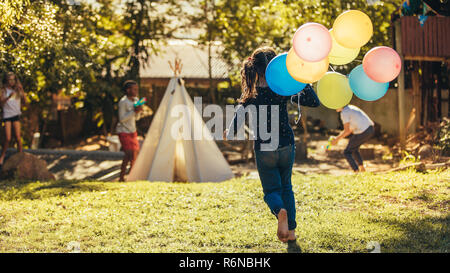 Bambina con palloncini colorati che corre verso i bambini a giocare in cortile. Bambini divertirsi all'esterno. Foto Stock
