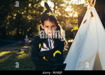 Carino piccolo ragazza in costume di procedura guidata che indossa il cappello con le braccia incrociate. Carino giovane bambina in abito di halloween in piedi in giardino nel cortile teepee in Foto Stock