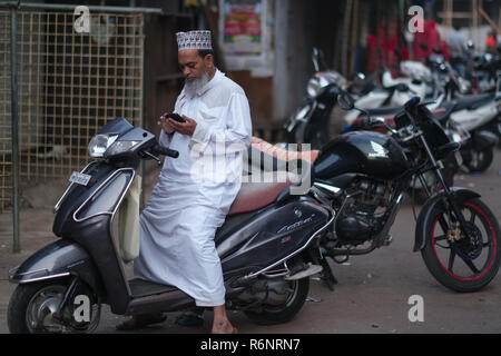 Un uomo musulmano in il musulmano dominato area di Bhendi Bazar in Mumbai, India, seduto su una motocicletta parcheggiata la lettura sul suo telefono cellulare Foto Stock