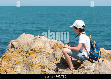 Un piccolo ragazzo si siede sul bordo di una scogliera ed esaminando la distanza dal mare. Foto Stock