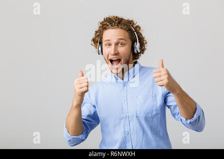 Ritratto di un felice giovane uomo che indossa shirt standing isolate su uno sfondo grigio, l'ascolto di musica con le cuffie, mostrando pollice in alto Foto Stock