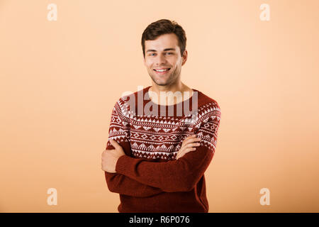 Immagine di uomo felice 20s con setole a maglia che indossa un maglione in posa sulla fotocamera con mani incrociate isolate su sfondo beige Foto Stock