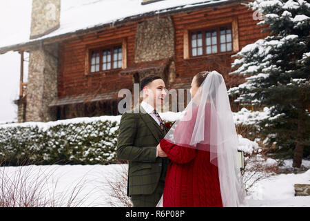 Carino elegante giovane sul giorno di nozze. Sposa e lo sposo si incontrano per la prima volta. Un primo sguardo. Matrimonio invernale sulla neve con un bel cottage di legno chalet su ski resort. Donna che indossa cardigan lavorato a maglia Foto Stock