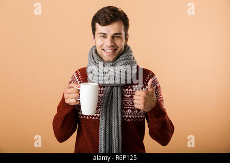 Ritratto di un allegro uomo vestito di maglione e sciarpa isolate su sfondo beige, tenendo tazza di tè caldo, mostrando pollice in alto Foto Stock
