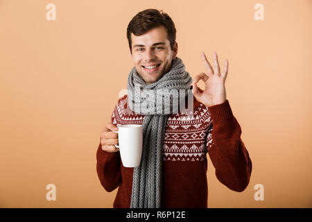 Ritratto di un allegro uomo vestito di maglione e sciarpa isolate su sfondo beige, tenendo tazza di tè caldo, mostrando il gesto ok Foto Stock