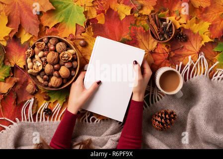 Autunno laici piana. Donna mani golding il libro bianco, ciotola di legno di dadi, tazza di caffè, il cono, cannella sopra beige plaid, colorato. Hygge e accogliente Foto Stock