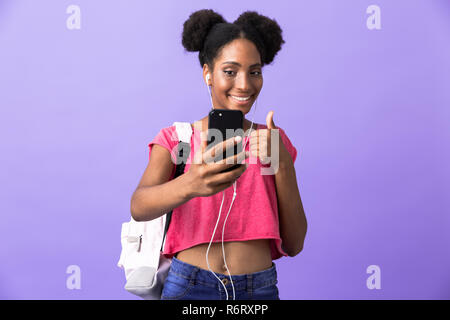 Foto di divertenti americano africano studente donna indossa uno zaino e cuffie bianche tramite telefono cellulare isolato su sfondo viola Foto Stock